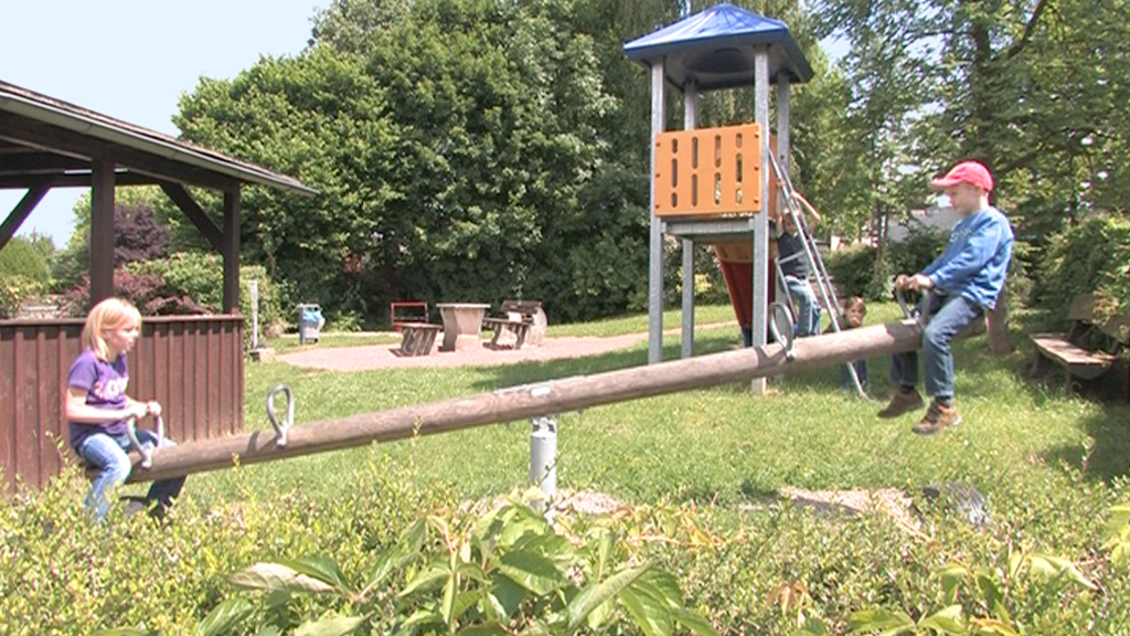 Zwei Kinder auf einer Wippe im Spielplatz, ein Junge mit roter Mütze und ein Mädchen mit blonden Haaren.