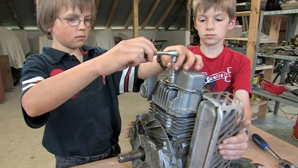 Zwei Jungen arbeiten gemeinsam an einem Motor, einer hält einen Schraubenschlüssel, der andere schaut konzentriert.