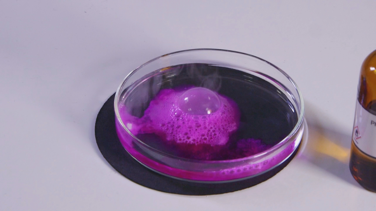 Eine Glasplatte mit einer violetten Flüssigkeit, die Blasen bildet und dampft, daneben eine Flasche mit einer chemischen Substanz.