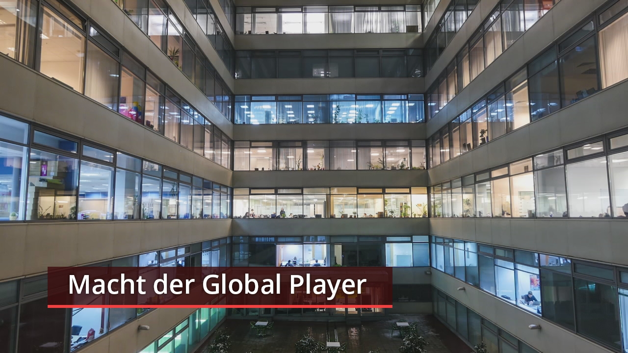 Blick auf ein modernes Bürogebäude mit großen Fenstern, in dem Mitarbeiter sichtbar sind. Text: 'Macht der Global Player'