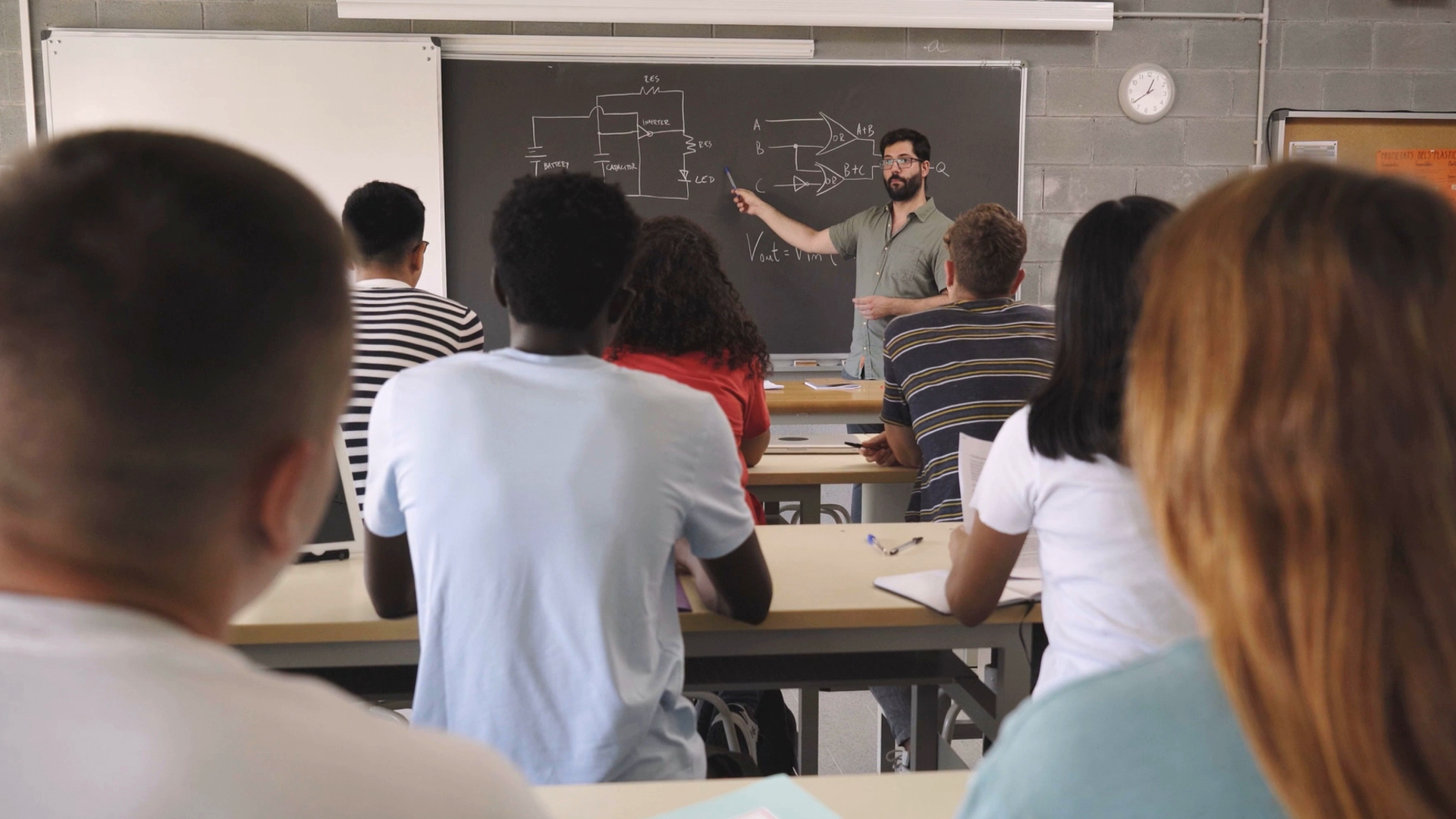 Lehrkraft zeigt auf Tafel mit Schaltplänen, Studierende sitzen an Tischen und hören zu.