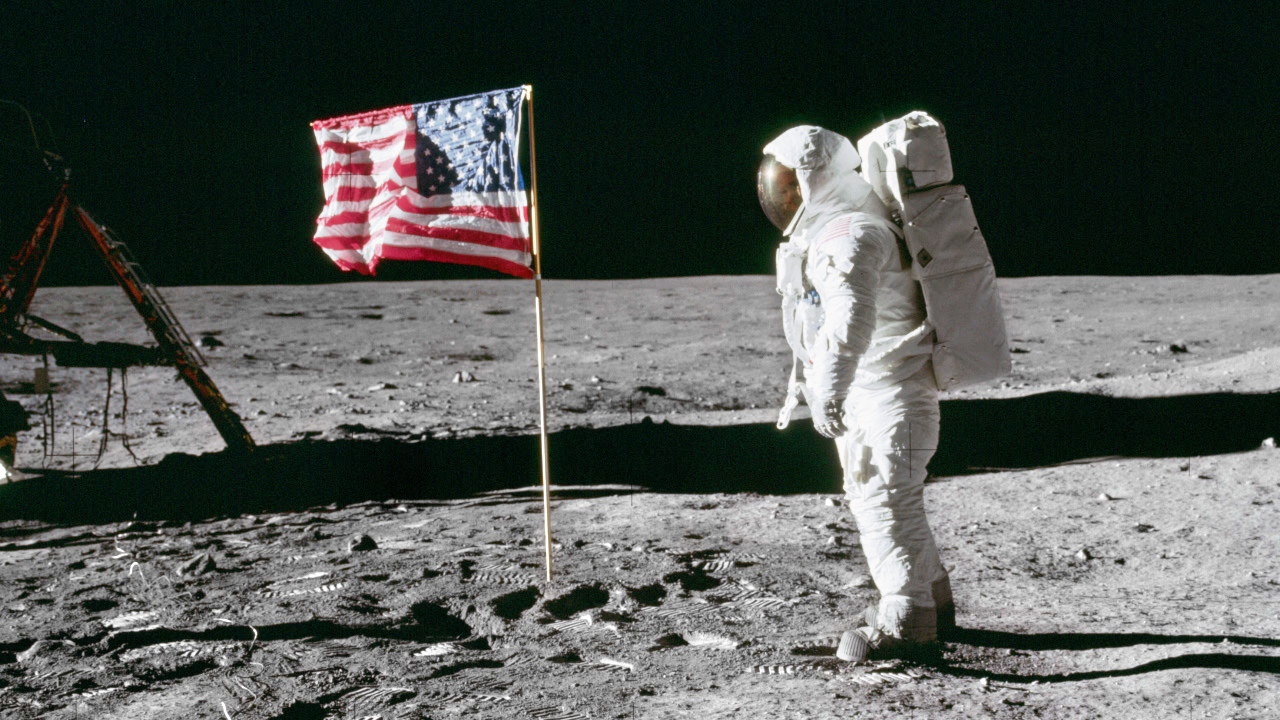 Ein Astronaut steht auf der Mondoberfläche neben einer US-Flagge, mit der Mondlandefähre im Hintergrund.