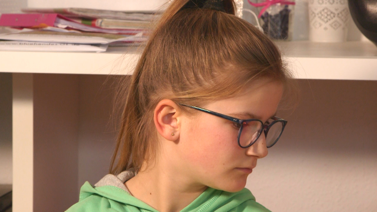 Ein Mädchen mit Brille und einem Zopf, das nachdenklich zur Seite schaut. Sie trägt einen grünen Hoodie.
