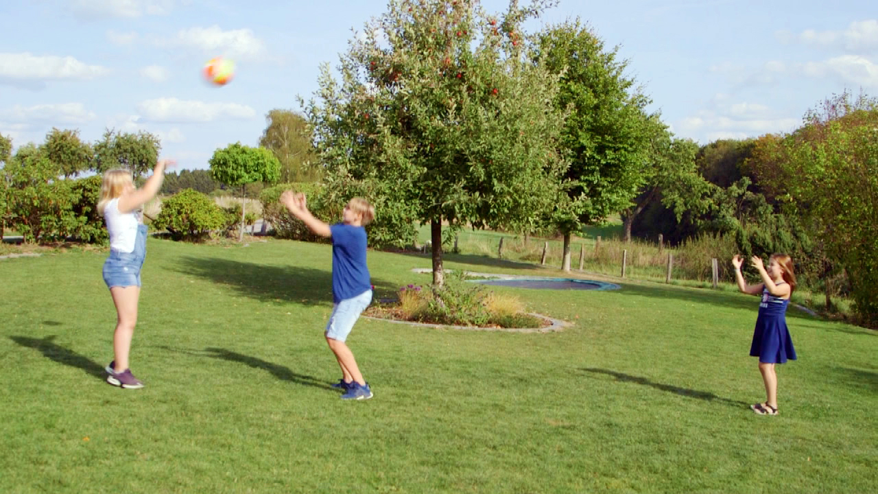 Drei Kinder spielen im Freien mit einem bunten Ball auf einer Wiese.