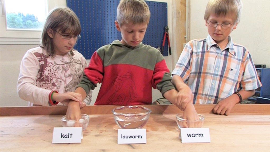 Drei Kinder testen Wassertemperaturen in Schalen mit den Beschriftungen 'kalt', 'lauwarm' und 'warm'.