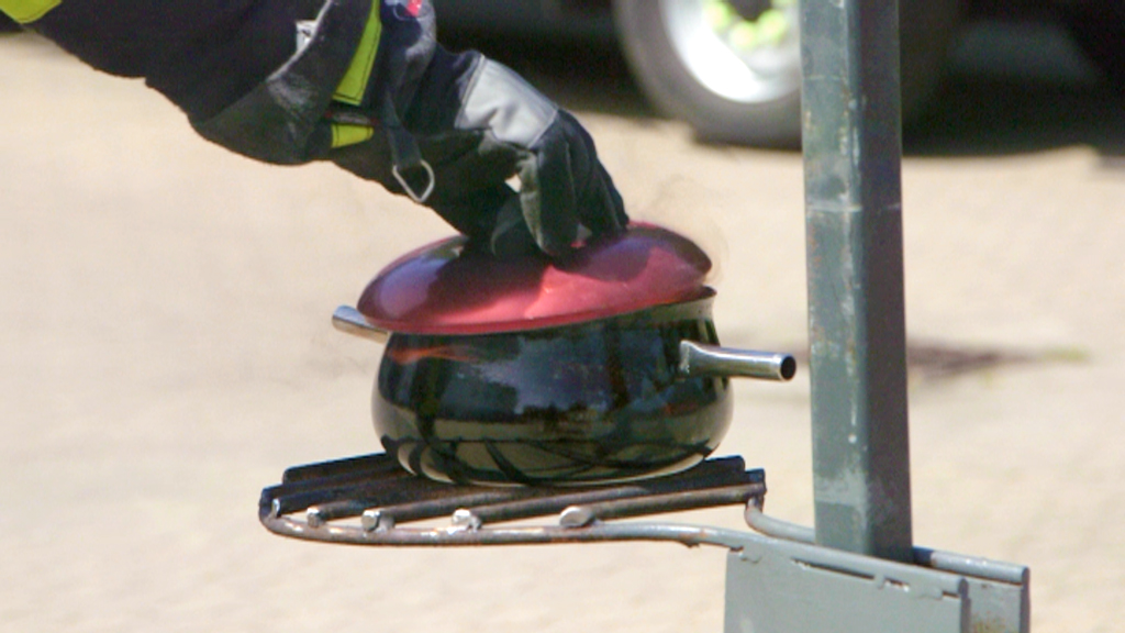 Ein Feuerwehrmann hebt den Deckel eines schwarzen Kochtopfs mit rotem Deckel an, der auf einem Gestell steht.