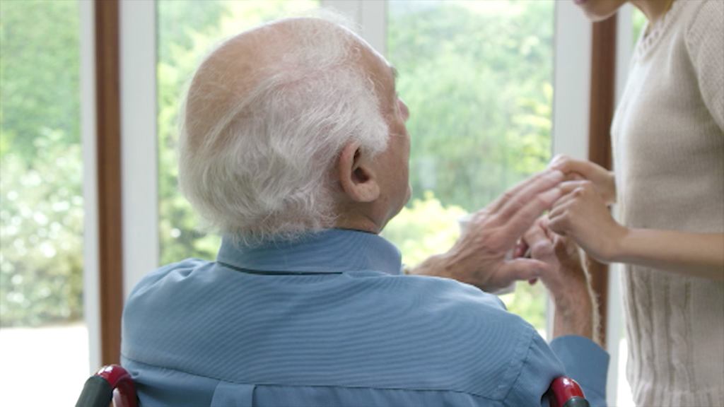Ein älterer Mann im Rollstuhl, der von einer jüngeren Person, die ihm gegenübersteht, die Hände hält.