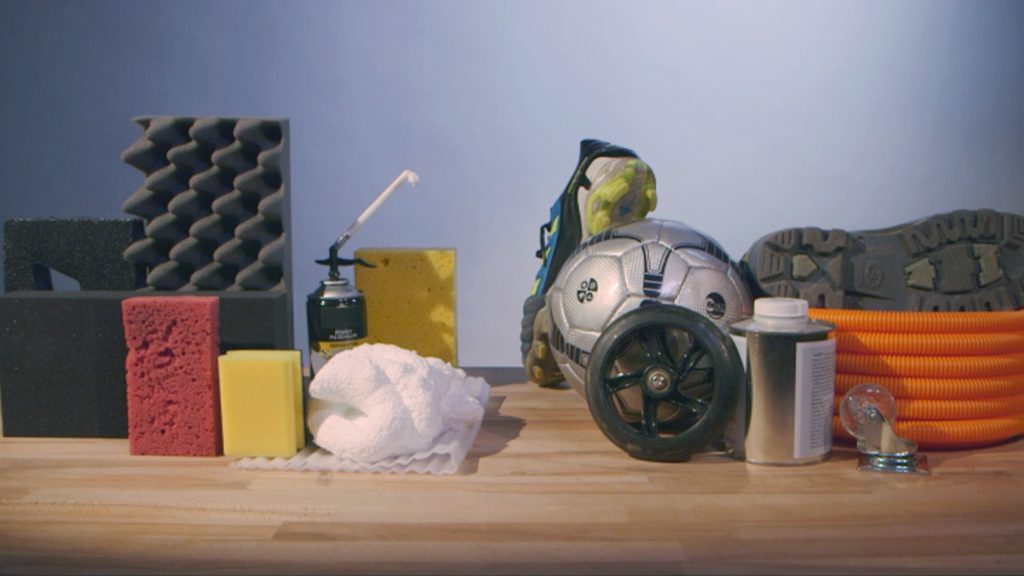 Verschiedene Materialien und Werkzeuge auf einem Tisch, darunter Schaumstoff, eine Fußball, eine Sprühdose und ein Schlauch.