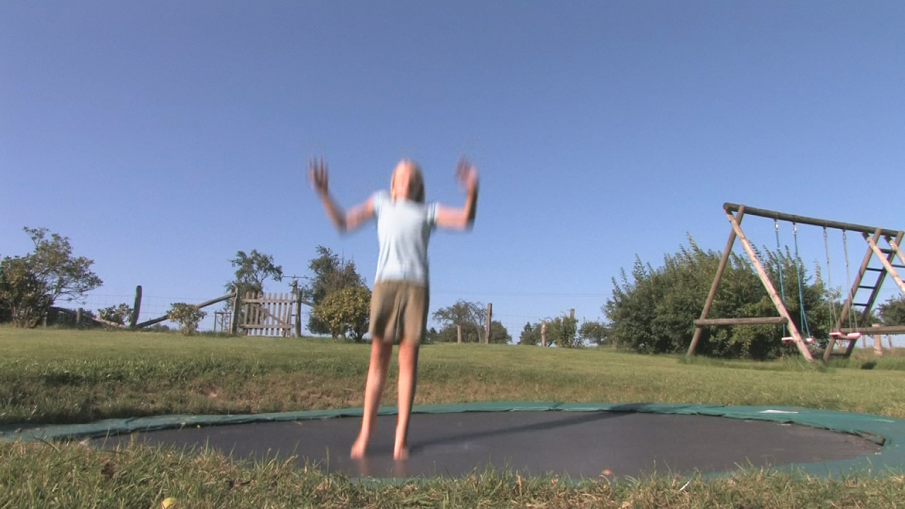 Ein Kind springt auf einem Trampolin im Freien, umgeben von Gras und Bäumen.