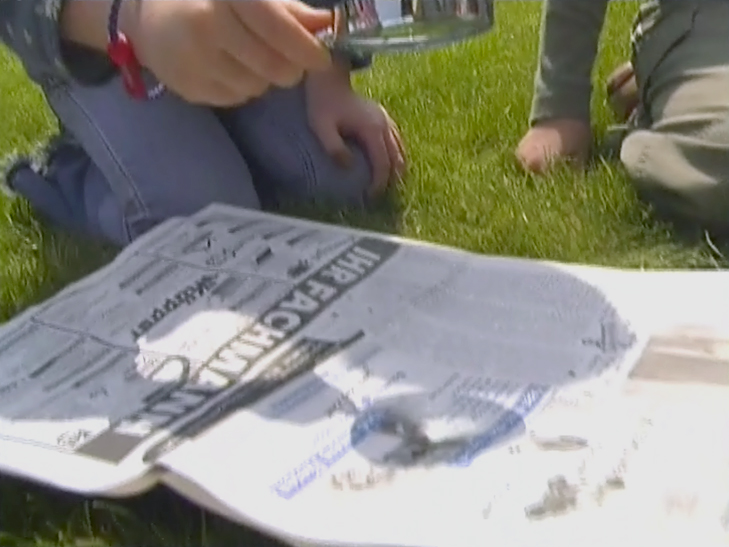 Eine Hand hält eine Tasse über eine Zeitung, die auf dem Gras liegt. Die Zeitung zeigt den Titel 'Information'.