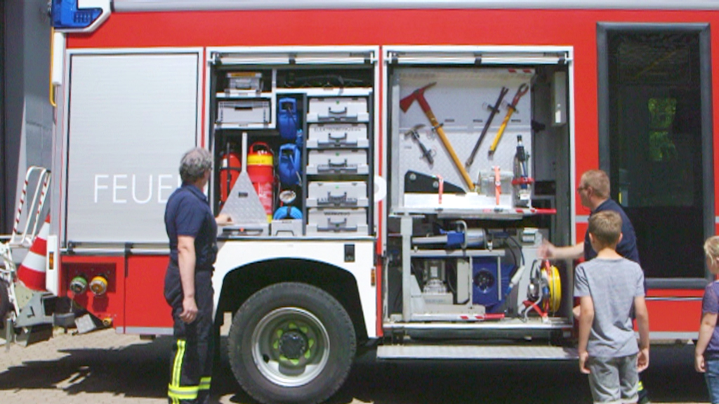 Feuerwehrmann zeigt Ausrüstung im offenen Schrank eines Feuerwehrautos, Kinder beobachten.