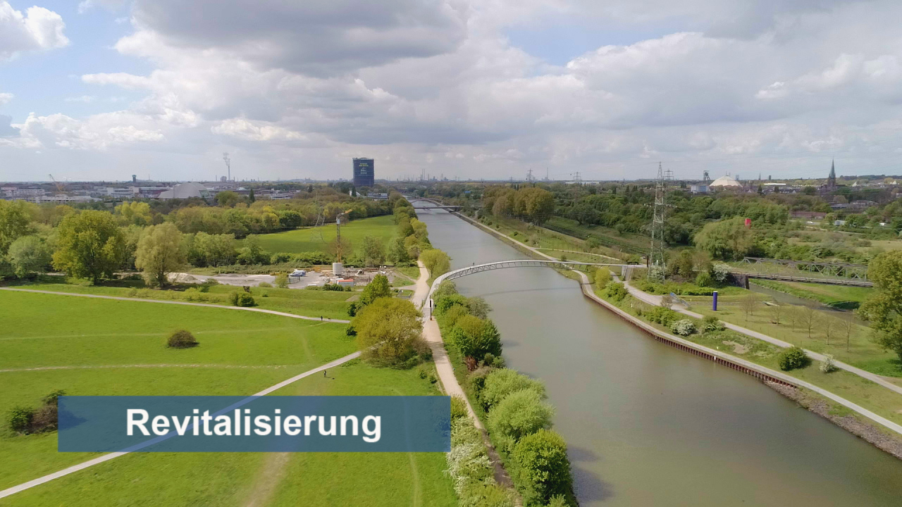 Luftaufnahme eines Flusses mit Grünflächen und einem Fußweg, im Hintergrund eine Stadtlandschaft unter bewölktem Himmel.