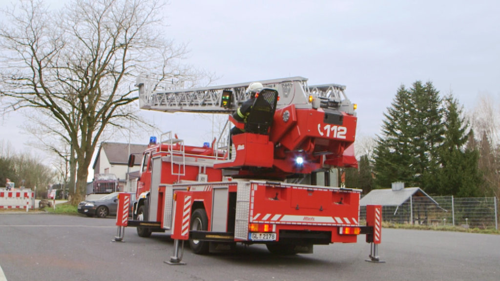 Feuerwehrfahrzeug mit Drehleiter auf einer Straße, Feuerwehrmann in Schutzkleidung sitzt auf der Leiter.