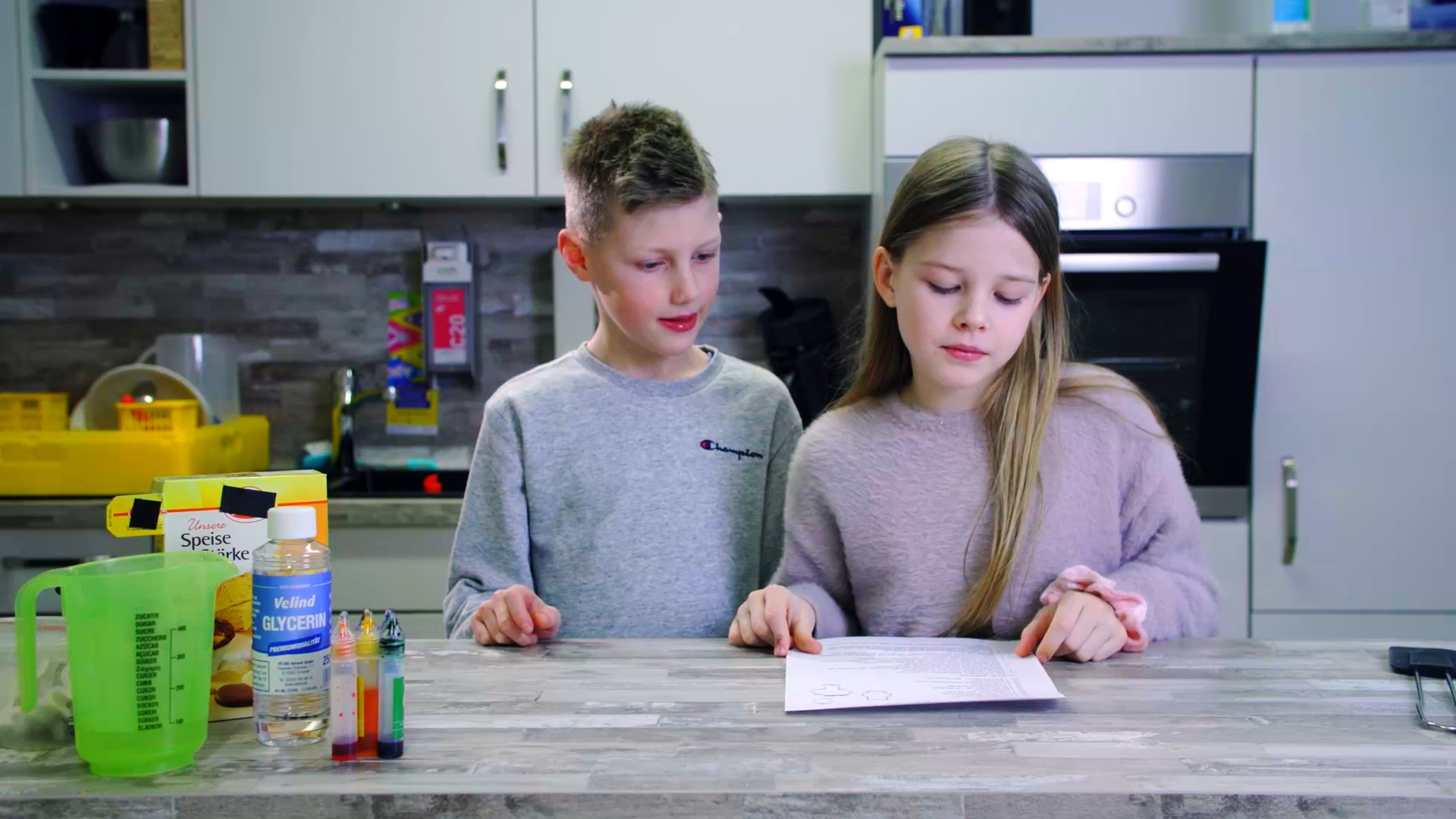 Zwei Kinder stehen an einem Küchentresen mit Messbecher, Flasche, Reagenzgläsern und einem aufgeschlagenen Buch