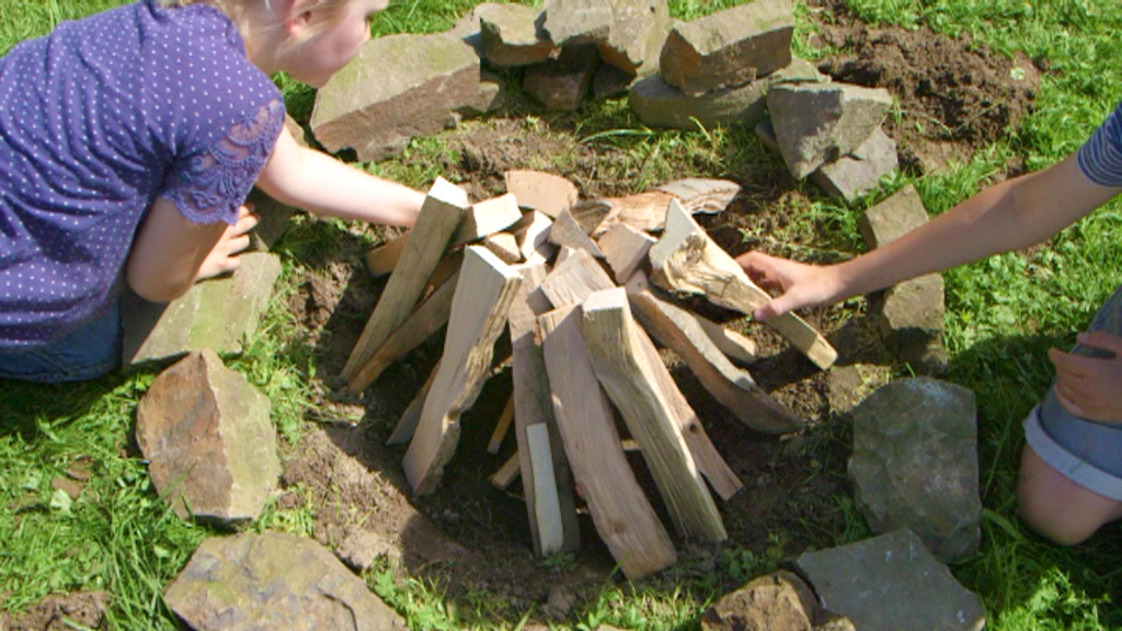 Ein Kind und ein Erwachsener bauen ein Lagerfeuer aus Holzscheiten in einem Steinkreis.