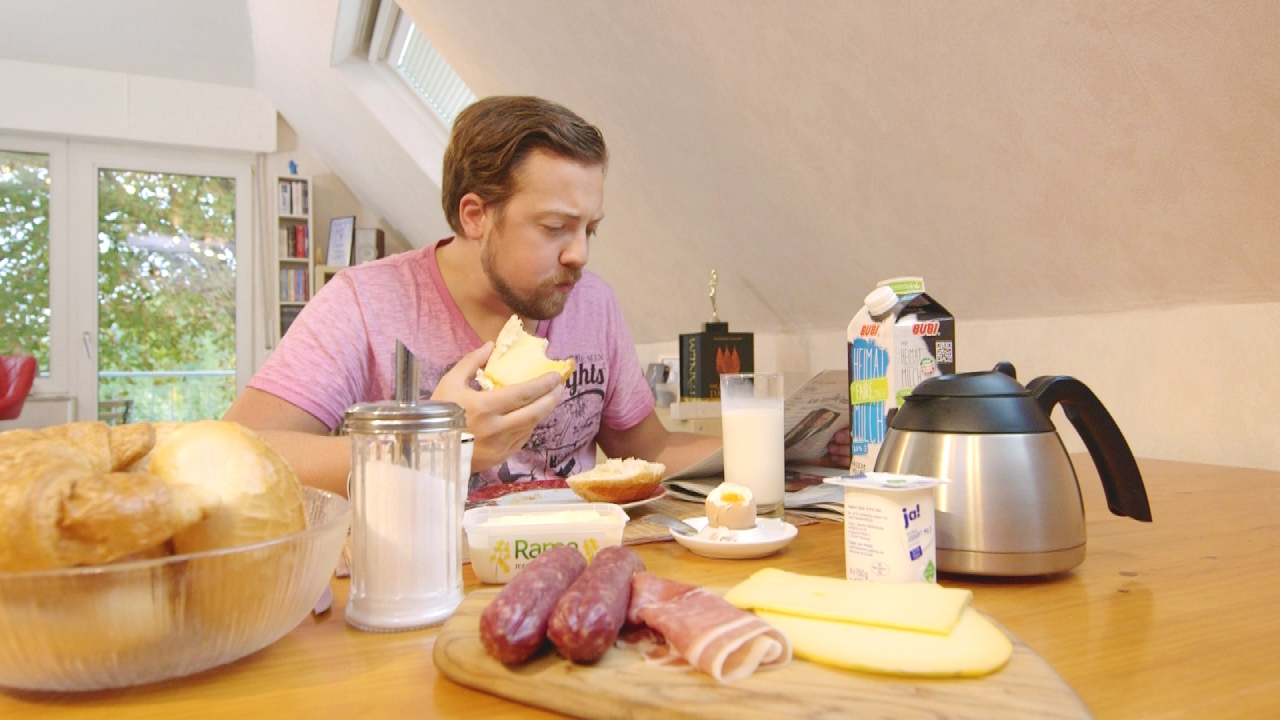 Mann mit Bart isst Frühstück am Tisch, umgeben von Brot, Wurst, Käse und Getränken.