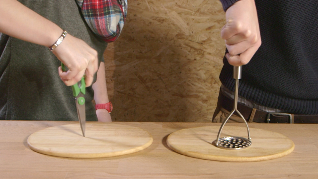 Eine Person hält eine Schere, während eine andere einen Kartoffelstampfer auf einem Holzbrett benutzt.