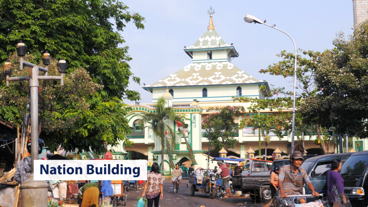 Blick auf eine belebte Straße mit einem markanten Gebäude im Hintergrund, umgeben von Menschen und Marktständen.