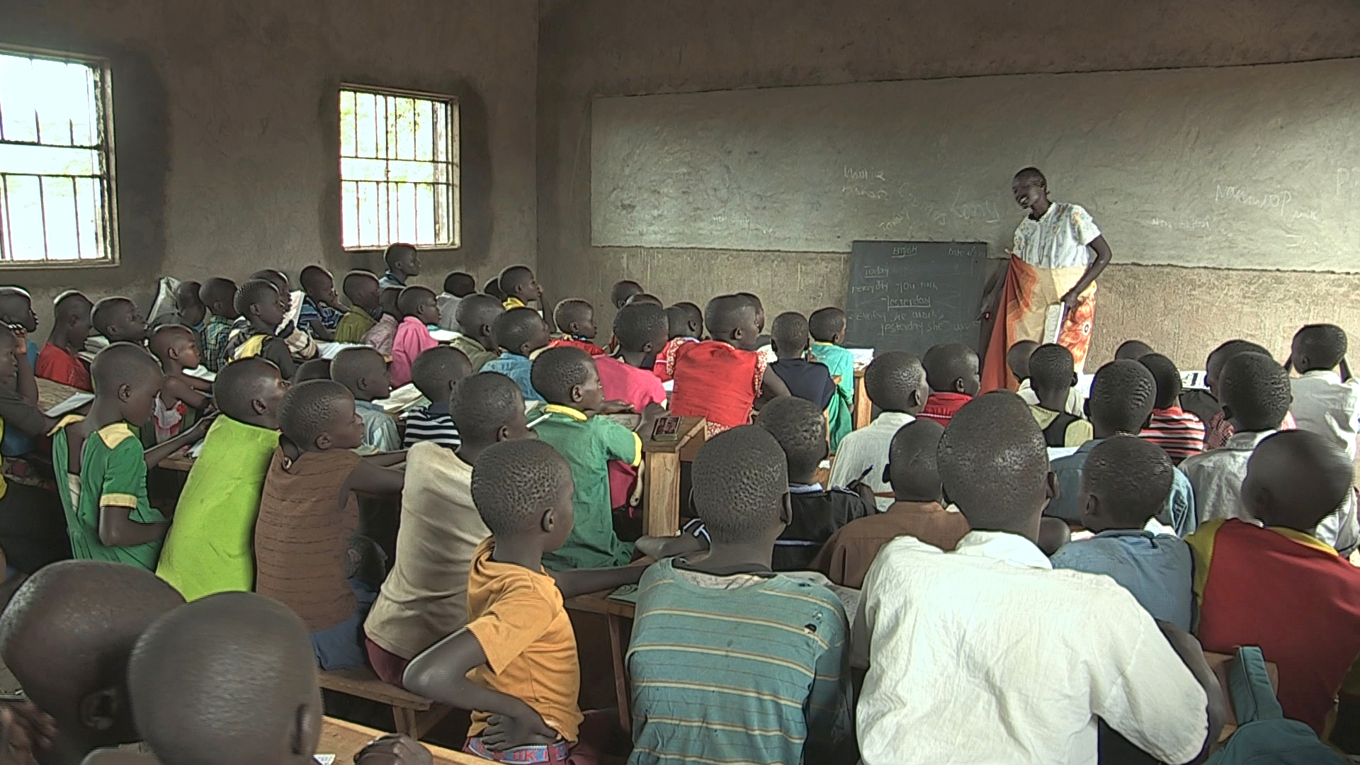 Lehrer steht vor Tafel in einfachem Klassenraum, viele Schüler sitzen auf Holzbänken und hören zu.
