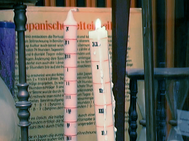 Zwei Kerzen mit Zahlenmarkierungen, eine rosa und eine weiße, vor einem Hintergrund mit Text.