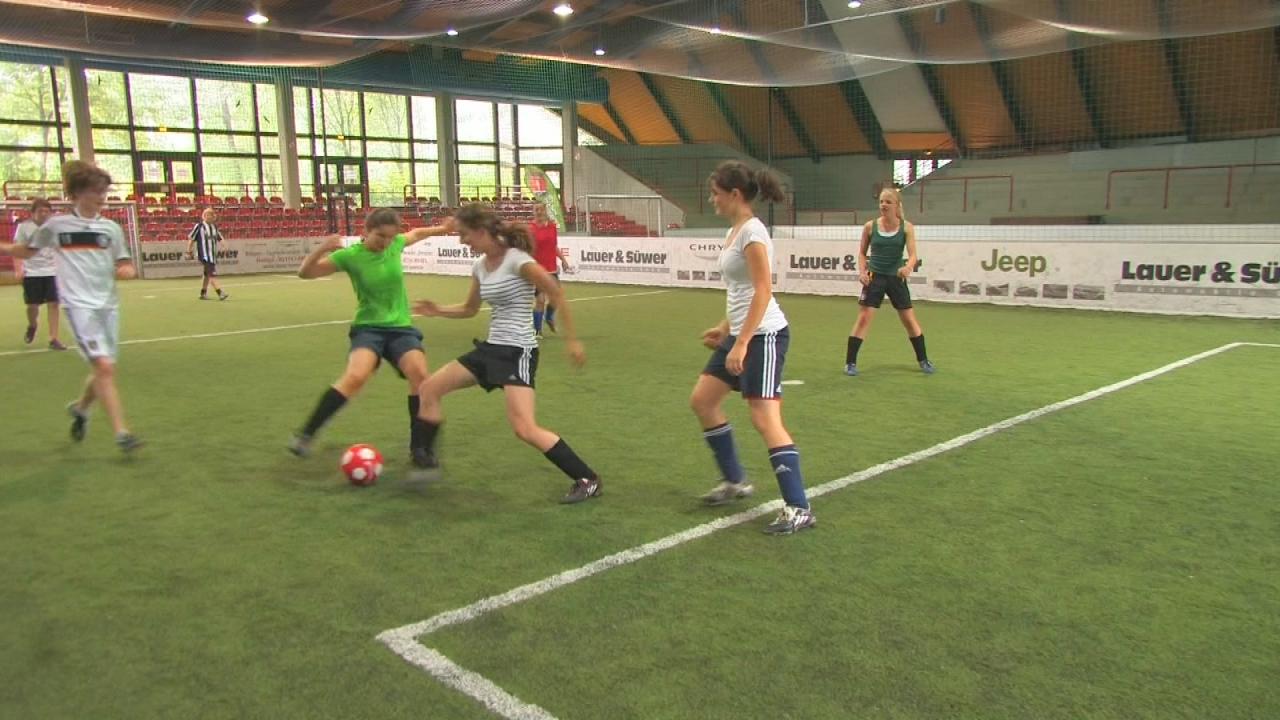 Mädchen spielen Fußball in einer Halle, eine Spielerin dribbelt den Ball, während andere versuchen, ihn zu erobern.
