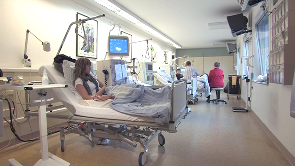 Eine Patientin liegt auf einem Krankenhausbett in einem modernen Krankenzimmer, während Pflegepersonal im Hintergrund arbeitet.