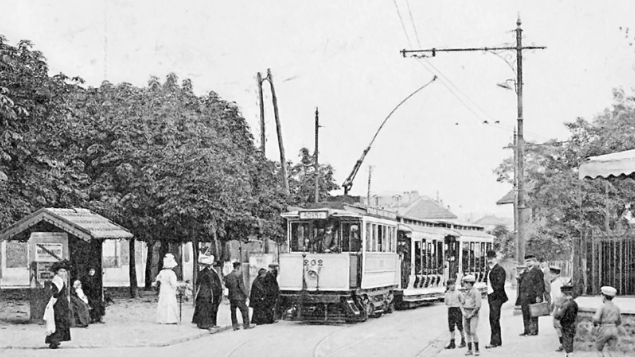 Historische Straßenbahn an einer Haltestelle mit wartenden Passagieren, umgeben von Bäumen und alten Gebäuden.