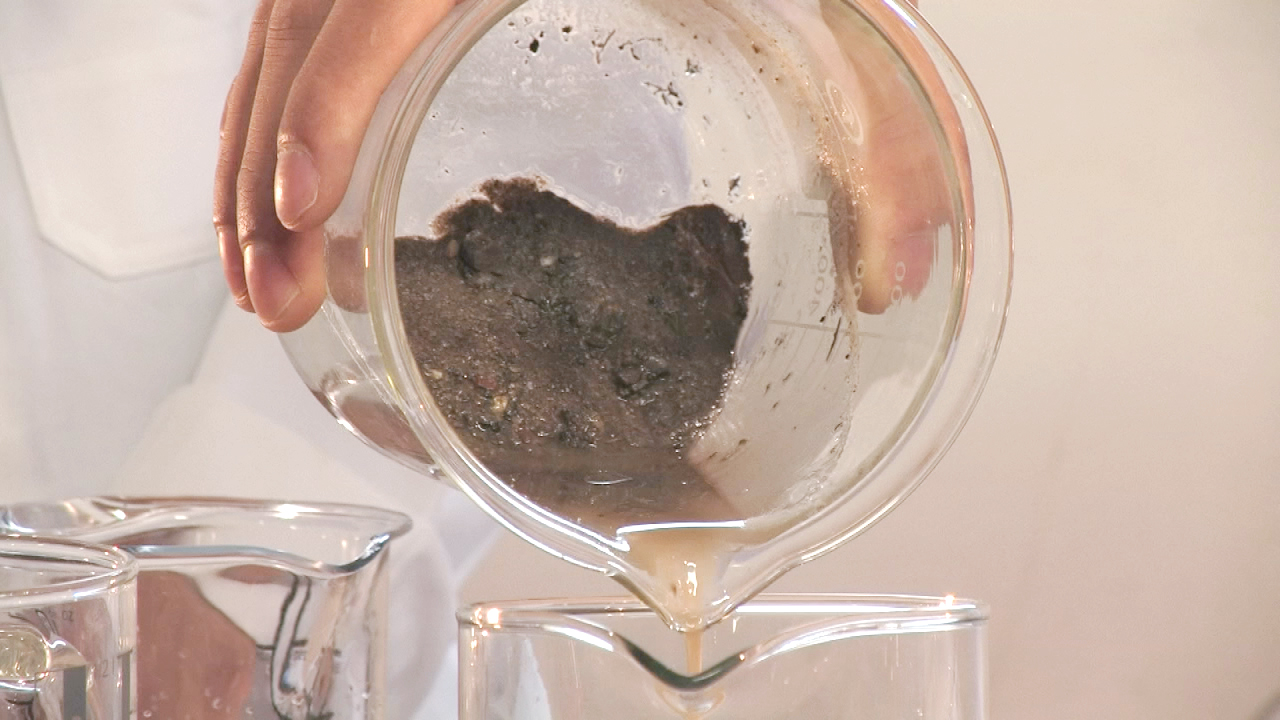 Eine Person gießt eine Flüssigkeit mit festen Rückständen aus einem Glas in einen anderen Behälter.