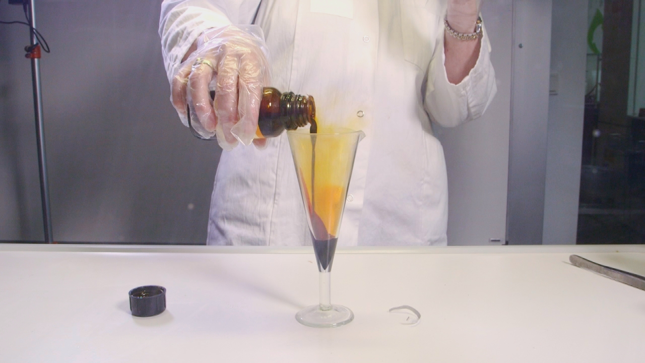 Eine Person in einem weißen Laborkittel gießt eine orangefarbene Flüssigkeit aus einer braunen Flasche in ein Glas.