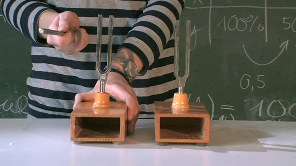 Zwei Holzblöcke mit Stimmgabeln, eine wird mit einem Hammer geschlagen, im Hintergrund Mathematikformeln.