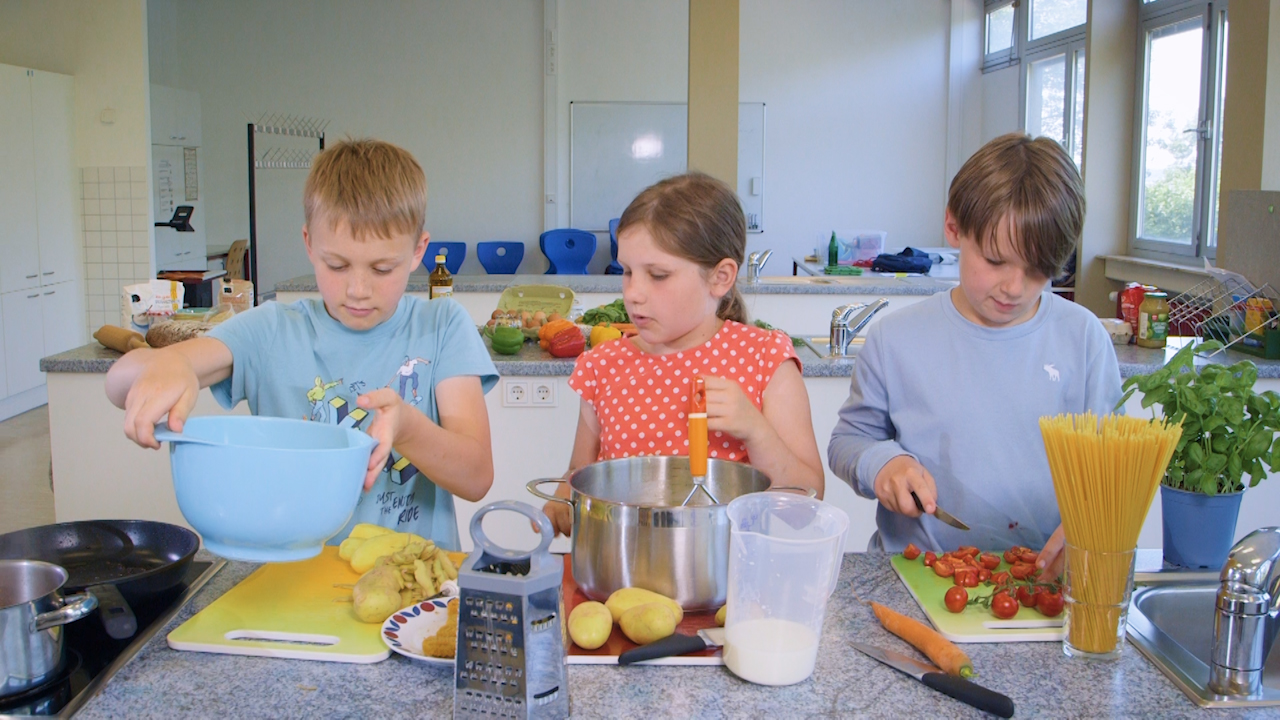 Drei Kinder bereiten gemeinsam eine Mahlzeit in einer modernen Küche vor, mit Gemüse und Kochutensilien auf der Arbeitsfläche.