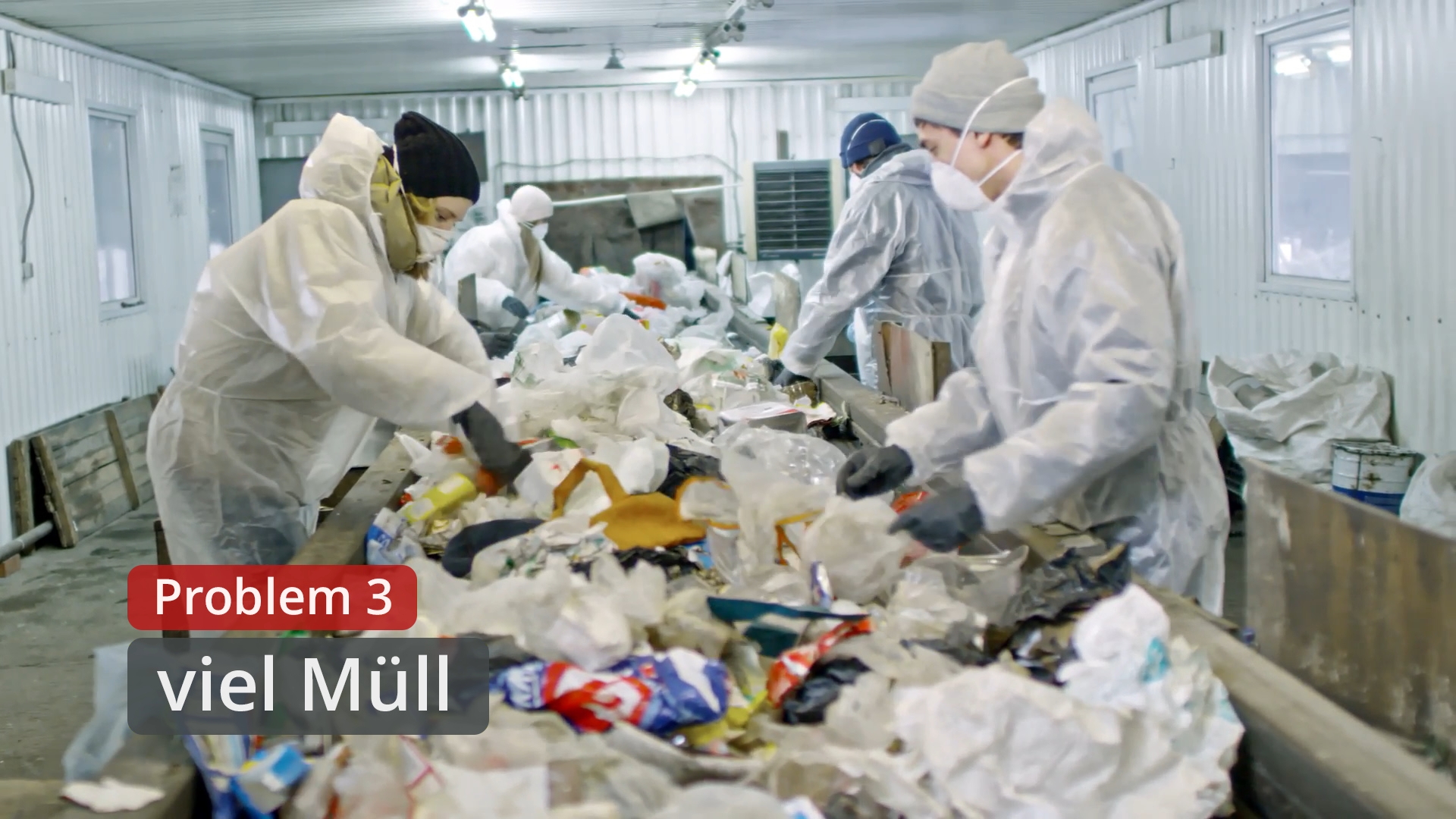 Mehrere Personen in Schutzanzügen sortieren Müll auf einem Förderband in einer Halle