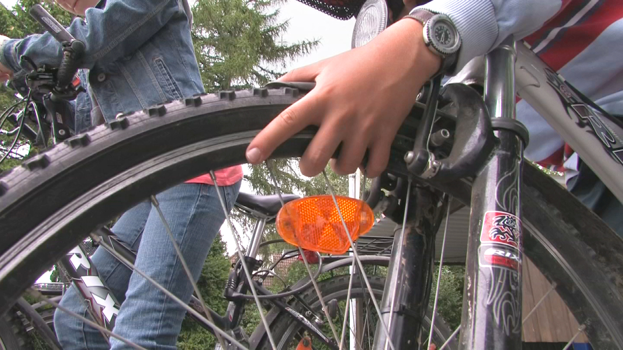 Eine Hand überprüft das Hinterrad eines Fahrrads, während ein weiteres Fahrrad im Hintergrund zu sehen ist.