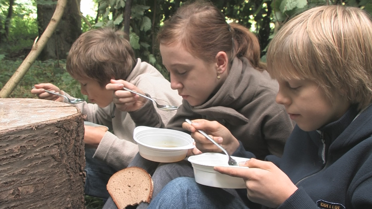 Drei Kinder sitzen im Wald und essen aus Schalen. Sie halten Löffel und konzentrieren sich auf ihr Essen.