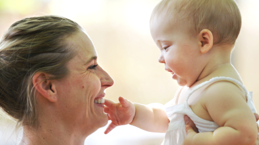 Eine lächelnde Mutter hält ihr Baby, das sie anlächelt und mit der Hand berührt.